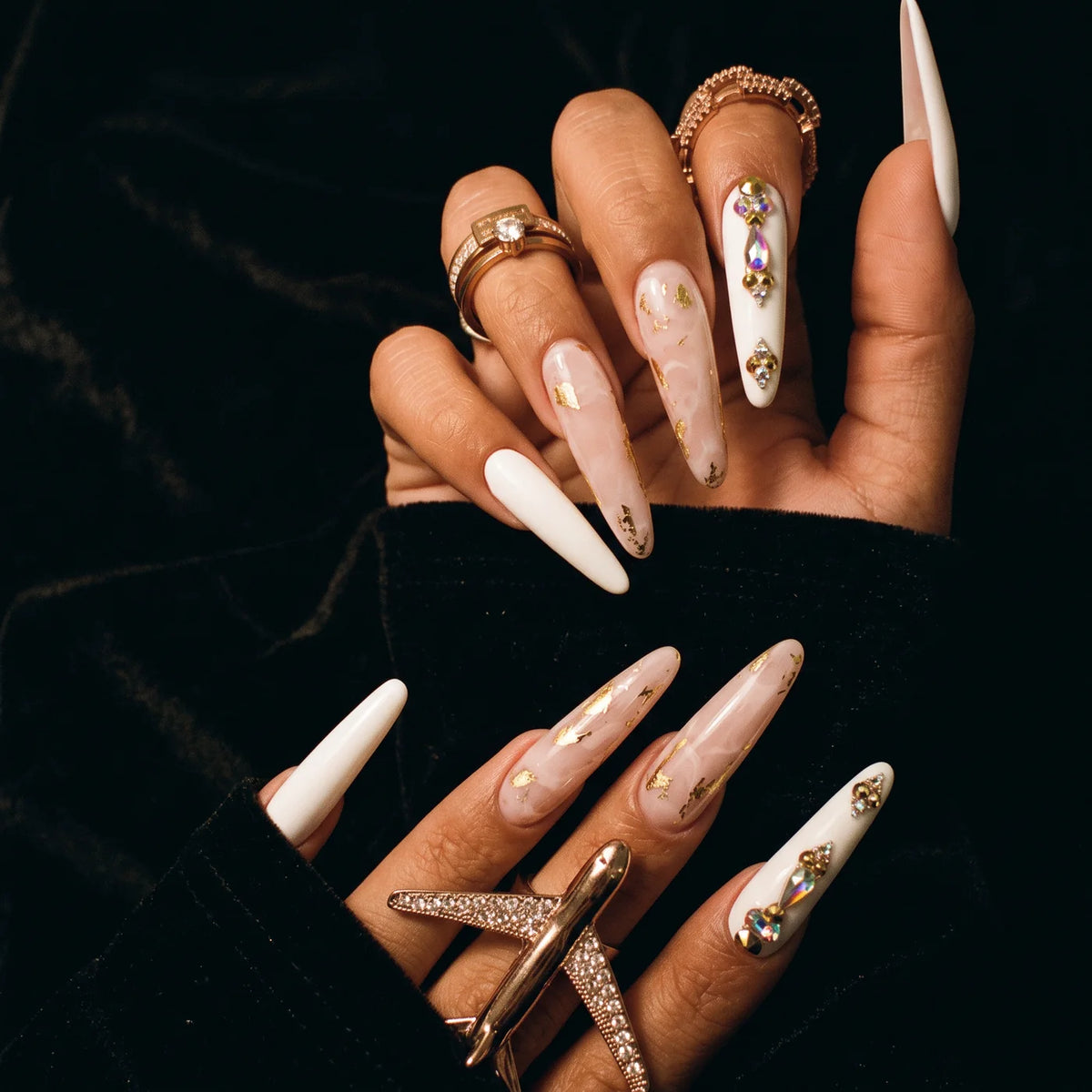 Eniko's hands with long white and gold leaf nails and rose gold rings on black velvet